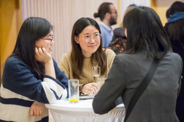Drei asiatische Frauen stehen am Tisch und unterhalten sich / Englisch:Three Asian women are standing at the table and talking