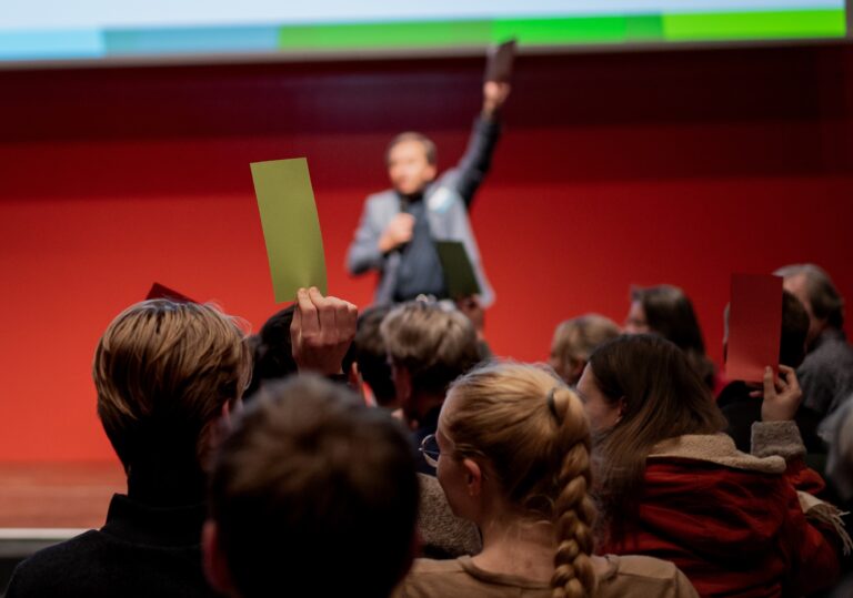 Eine Person steht vor einem Publikum auf der Bühne, Personen im Publikum halten farbige Blätter nach oben.