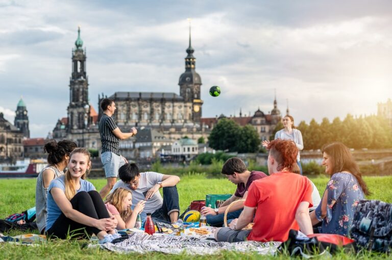 Junge Menschen sitzen mit einem Lächeln zusammen auf einer Wiese vor der Stadtkulisse von Dresden.