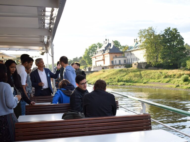 Im Vordergrund Personen, die sich auf einem Boot unterhalten. Im Hintergrund ein Schloss.