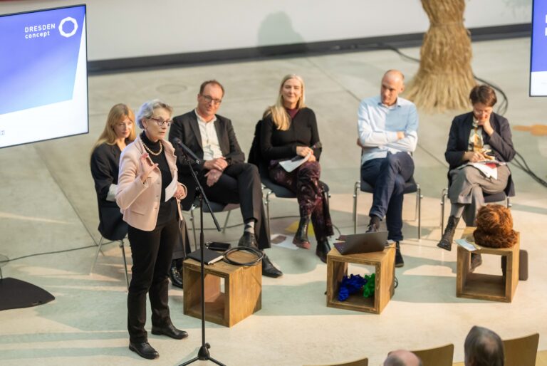 Auf einer Bühne sitzen 5 Personen. Im Vordergrund ist Ursula M. Staudinger, die stehend in ein Mikrofon spicht und das Publikum anblickt. Auf der Bühne sind zudem Hocker, auf welchen neben Notizbüchern und Notebooks auch haarige Plüschgegenstände sowie blaue und grüne Exponate liegen.