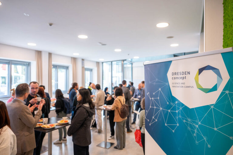 Eine große Gruppe Menschen steht in einem Saal mit Stehtischen auf denen Tassen und Gebäck steht. Im Vordergrund ist eine Grafik mit de Logo DRESDEN-concept Science and Innovation Campus zu sehen.