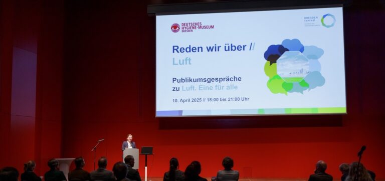 Eine Person steht auf der Bühne vor Publikum. In Hintergrund ist ein großer Bildschirm mit den Logos des Deutschen-Hygiene-Museums Dresden und DRESDEN-concept Science and Innovation Campus, darunter die Aufschrift Reden wir über // Luft, Publikumsgespräche zu Luft. Eine für alle, 10. April 2025, 18:00 bis 21:00 Uhr, daneben ist ein Bild umringt von 8 Sprechblasen in unterschiedlichen Farben abgebildet. Logo