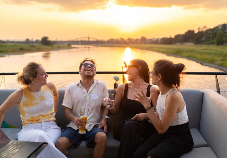 4 Personen sitzen zusammen an einer Sitzecke und lachen miteinander, im Hintergrund ist eine Brücke und ein Fluss bei Sonnenuntergang zu sehen. Logo