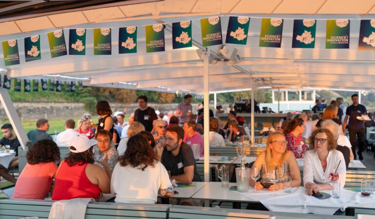 Auf einem Schiff sitzen viele Menschen zusammen an Tischen. Über ihnen hängt eine Fahnenkette mit zwei wechselnden Motiven. Das Eine zeigt die Stadtkarte von Dresden mit dem Logo von DRESDEN-concept und einigen bunten Fähnchen, das Andere die Aufschrft "Science and Innovation in collaboration". Logo