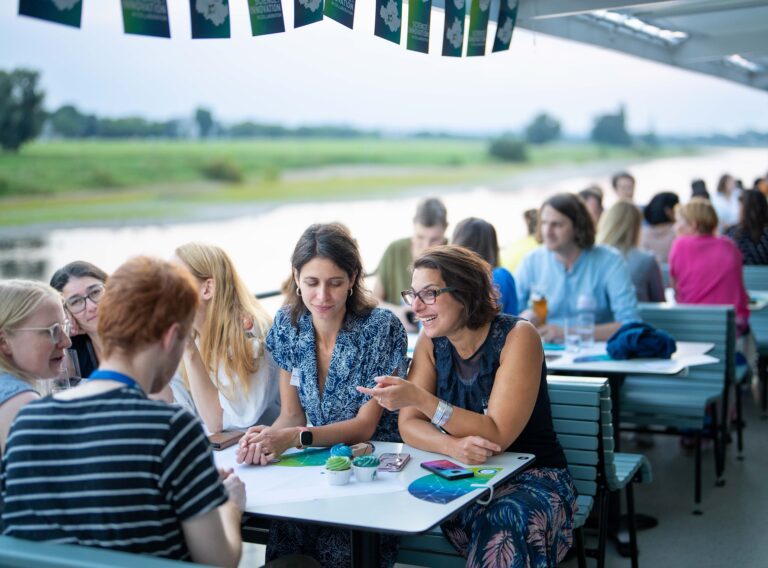 Auf einem Schiff sitzen viele Menschen zusammen an Tischen. Auf den Tischen stehen Cupcakes sowie Materialien mit dem Logo DRESDEN-concept aus. Logo