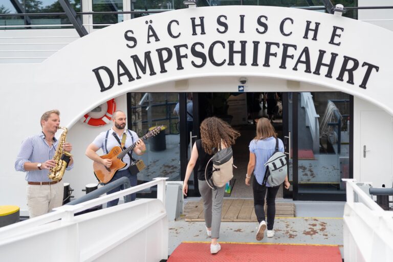 2 Personen laufen unter einem großen Schild mit der Aufschrift "Sächsische Dampfschifffahrt" hindurch, daneben stehen 2 Musiker. Logo