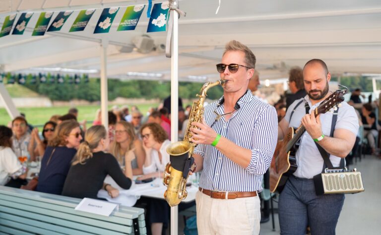 2 Personen spielen im Gehen Musik auf einem großen Schiff, eine Person spielt Saxophon und die andere Perspn spielt Gitarre. Logo