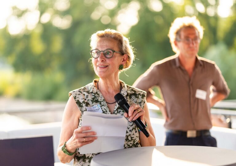 Eine Person hält ein Mikrofon und Zettel in der Hand und blickt weiter weg, diese Perspn trägt ein Namensschild mit der Aufschrift Ursula M. Staudinger, TUD/Vorstand DDc Logo