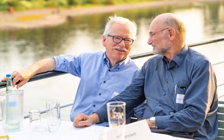 2 Personen unterhalten sich an einem Tisch, im Hintergrund ist ein Fluß zu sehen. Logo