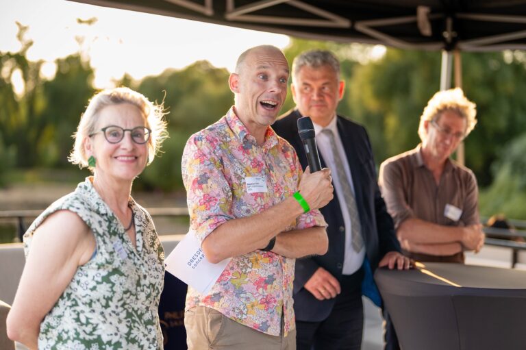4 Personen stehen nebeneinander, eine Person lächelt direkt in die Kameria, die Person daneben hält ein Mikrofon und Moderationskarten mit der Aufschrift DRESDEN-concept, diese Person trägt auch ein Namensschild mit Stephan Grill, MPI-CBG/Vorstand DDc Logo