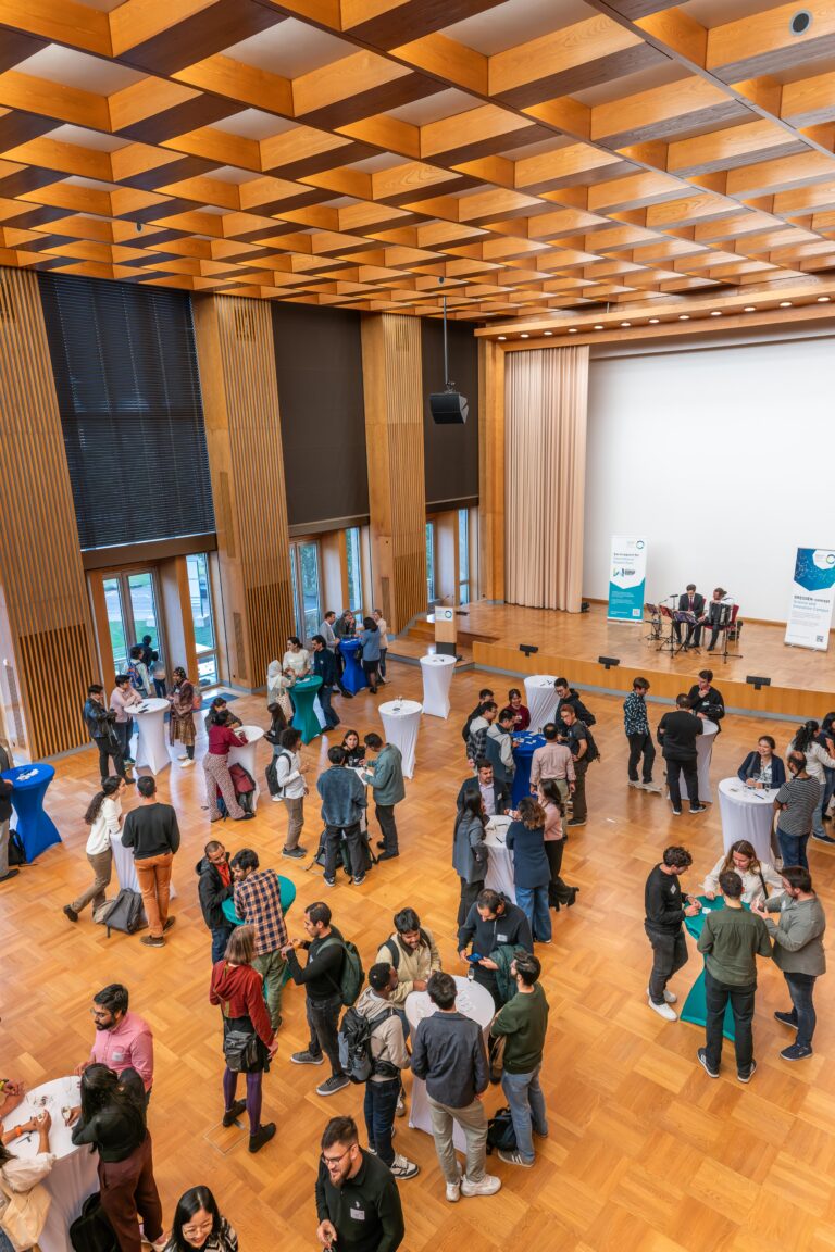 A crowd of people are standing at bar tables in a festive hall. Roll-ups from DRESDEN-concept are in the background, as well as two musicians on stage. Logo
