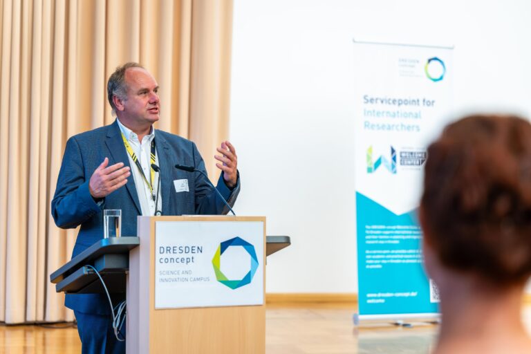 A person is standing at a standing desk. The DRESDEN-concept Science and Innovation Campus logo is displayed on the standing desk. In the background is a roll-up banner with the words “Servicepoint for International Researchers.” Logo