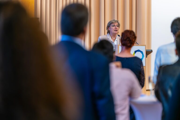 A person is standing at a standing desk. A crowd of people are looking at that person. Logo