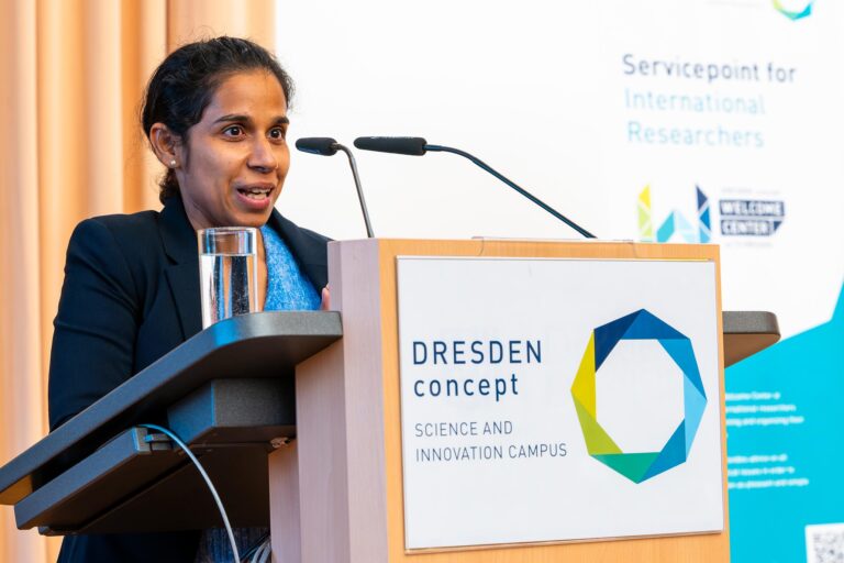 A person is standing at a standing desk. The DRESDEN-concept Science and Innovation Campus logo is displayed on the standing desk. Logo