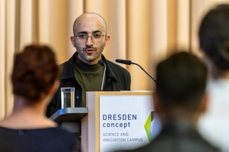 A person is standing at a standing desk. The DRESDEN-concept Science and Innovation Campus logo is displayed on the standing desk. Logo