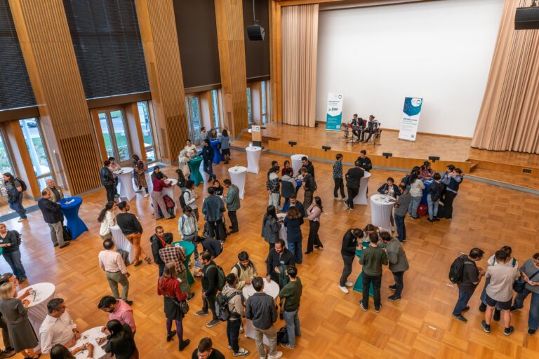 A crowd of people are standing at bar tables in a festive hall. Roll-ups from DRESDEN-concept are in the background, as well as two musicians on stage. Logo