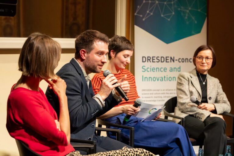 4 Personen sitzen auf einer Bühne. 2 von Ihnen halten Mikrofone in der Hand, eine Person redet. Im Hintergrund is ein Roll up von DRESDEN-concept zu sehen. Logo