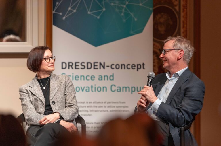 2 Personen sitzen auf einer Bühne und reden miteinander. Eine Person hält ein Mikrofon. Im Hintergrund is ein Roll up von DRESDEN-concept zu sehen. Logo