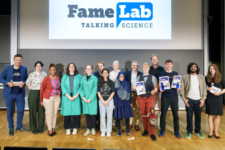 Gruppenbild von 15 Personen vor einer Tafel. Darüber ist das Logo von Fame Lab Talking Science angebracht. Logo